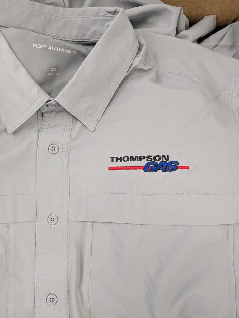 Close-up of a light gray Port Authority work shirt featuring the embroidered ThompsonGas logo on the left chest, representing custom branded industrial workwear.