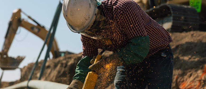 Construction worker in flame-resistant gear using a power tool with sparks flying on a job site.