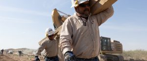 Construction worker in FR shirt carrying a heavy wooden beam across a dusty job site.