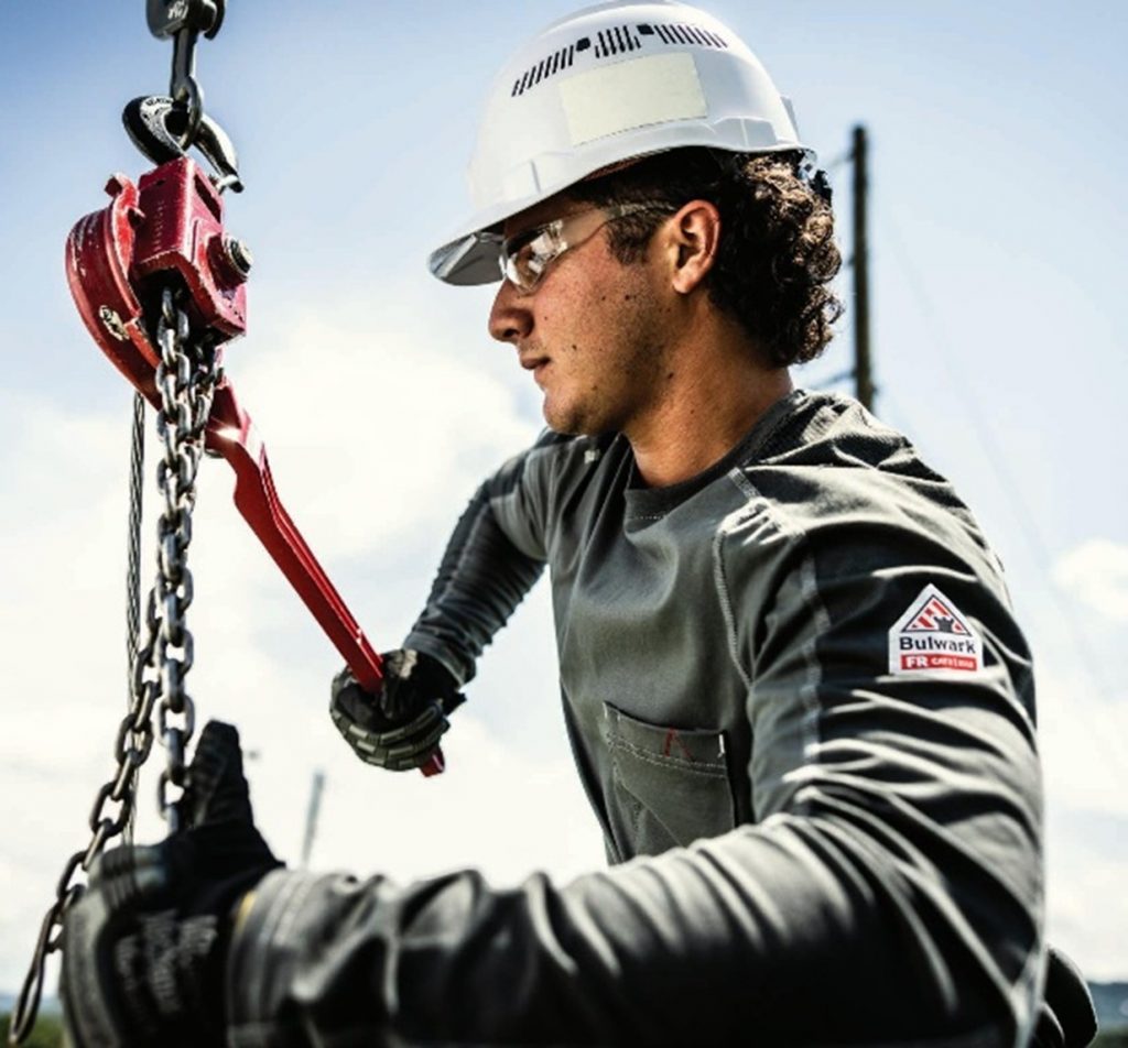 Oil rig worker wearing Bulwark FR gear and hard hat while adjusting heavy equipment. A close-up of an oilfield worker outfitted in Bulwark flame-resistant clothing, gloves, and PPE, highlighting industry-grade protection in hazardous, flammable environments. Ideal representation of how FR standards are applied in the energy and oil & gas sectors.