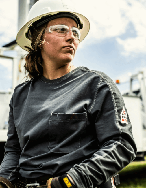 Female utility worker in a grey FR long-sleeve shirt, hard hat, and safety glasses.