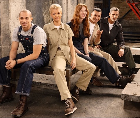 A diverse group of five workers sit together on an industrial bench wearing a mix of durable workwear—coveralls, khaki utility sets, and rugged boots—smiling in a relaxed, confident pose.