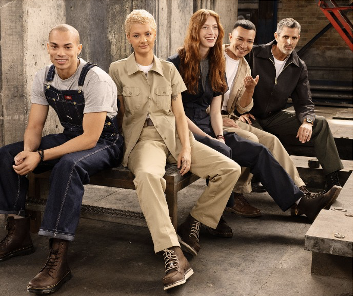 Group of workers wearing rugged workwear uniforms, including coveralls, overalls, and utility shirts, sitting together on benches in an industrial setting.