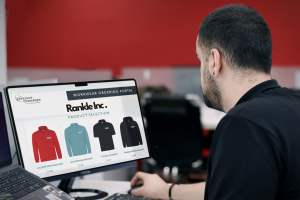 Man using a computer to browse the Work Hard Dress Right workwear ordering portal, displaying uniform options in red, light blue, navy, and black.