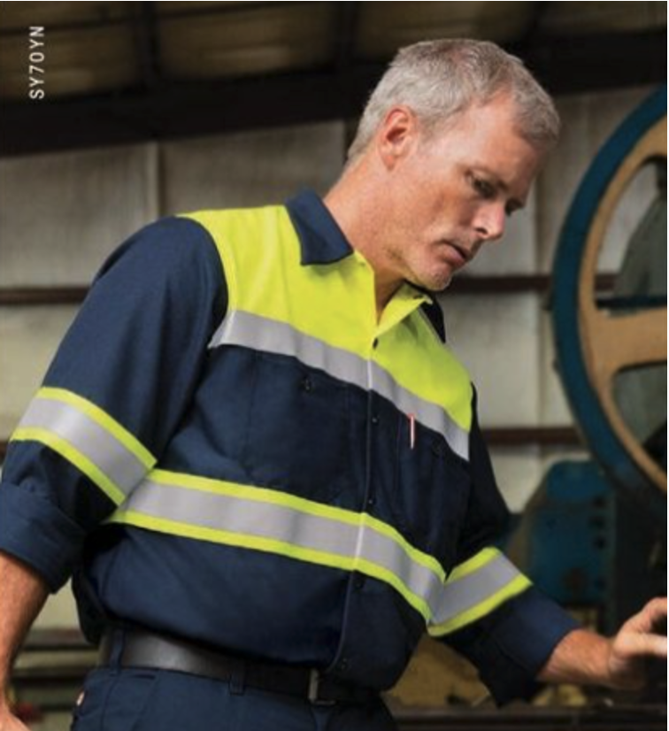 A man wearing a navy and high-visibility yellow work shirt with reflective striping operates equipment inside an industrial facility.