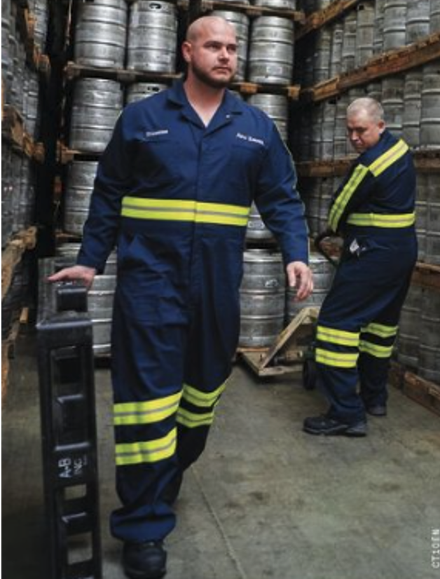 Two warehouse workers wearing navy coveralls with bright yellow reflective striping move kegs in an industrial storage area.