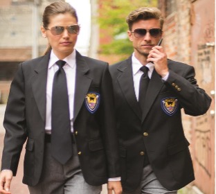 A man and woman wearing matching black blazers with embroidered security badges, white shirts, black ties, and sunglasses walk confidently outdoors, one speaking on a phone.