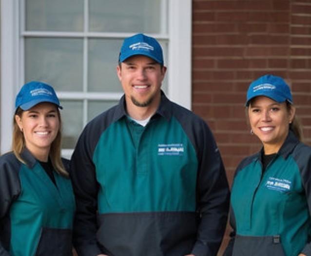 Three smiling employees wearing matching green and black uniforms with branded baseball caps, standing outside a brick building.