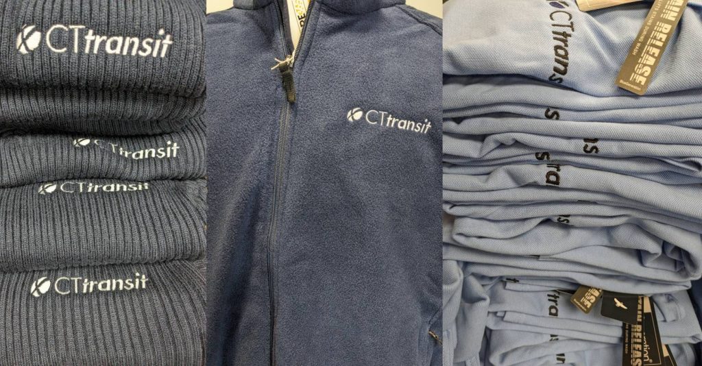 Three stacks of embroidered CTtransit apparel, including knit beanies, fleece jackets, and light blue polo shirts, all neatly folded and displaying the CTtransit logo.
