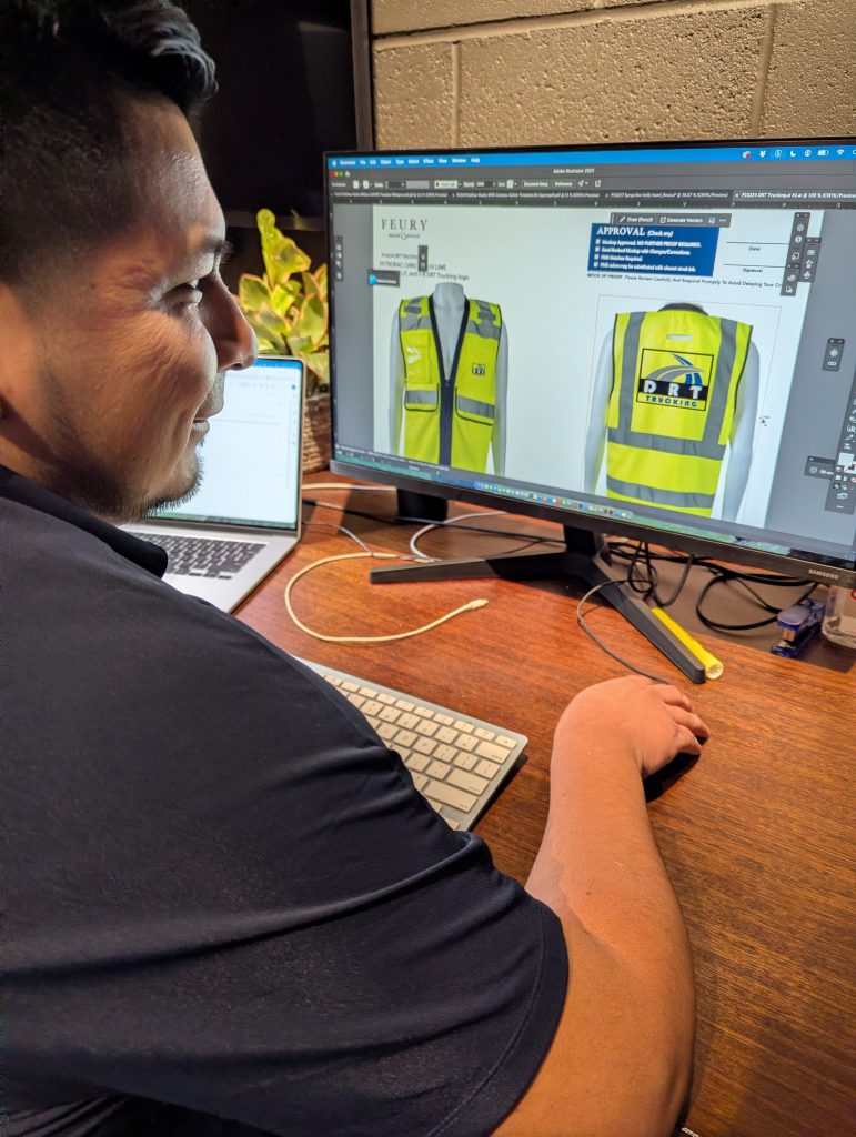 A graphic designer reviews a high-visibility safety vest proof on a computer screen at Feury Image Group. The design on the monitor shows front and back views of a yellow vest featuring the DRT Trucking logo, part of a branded apparel project in progress.