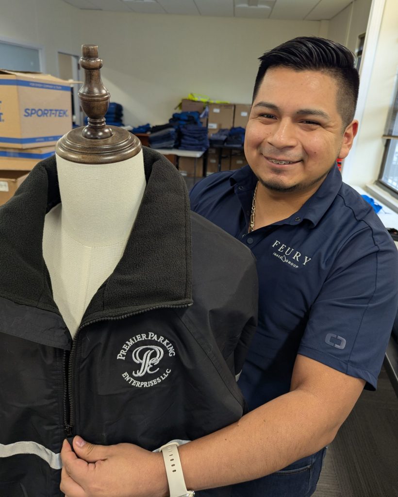 Feury Image Group team member presenting a black embroidered jacket featuring a company logo for corporate branded apparel.