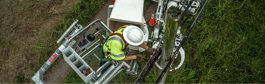 Utility worker in high-visibility FR clothing operates equipment from a bucket lift beside a power pole.