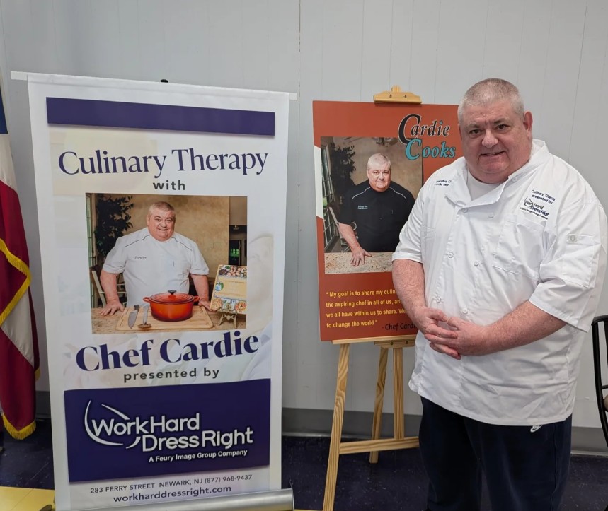 Chef Cardie Mortimer wearing a custom chef uniform standing beside Culinary Therapy banner and branded event signage